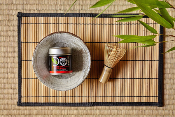 A bowl with a container of organic ceremonial matcha and a whisk on a bamboo mat.